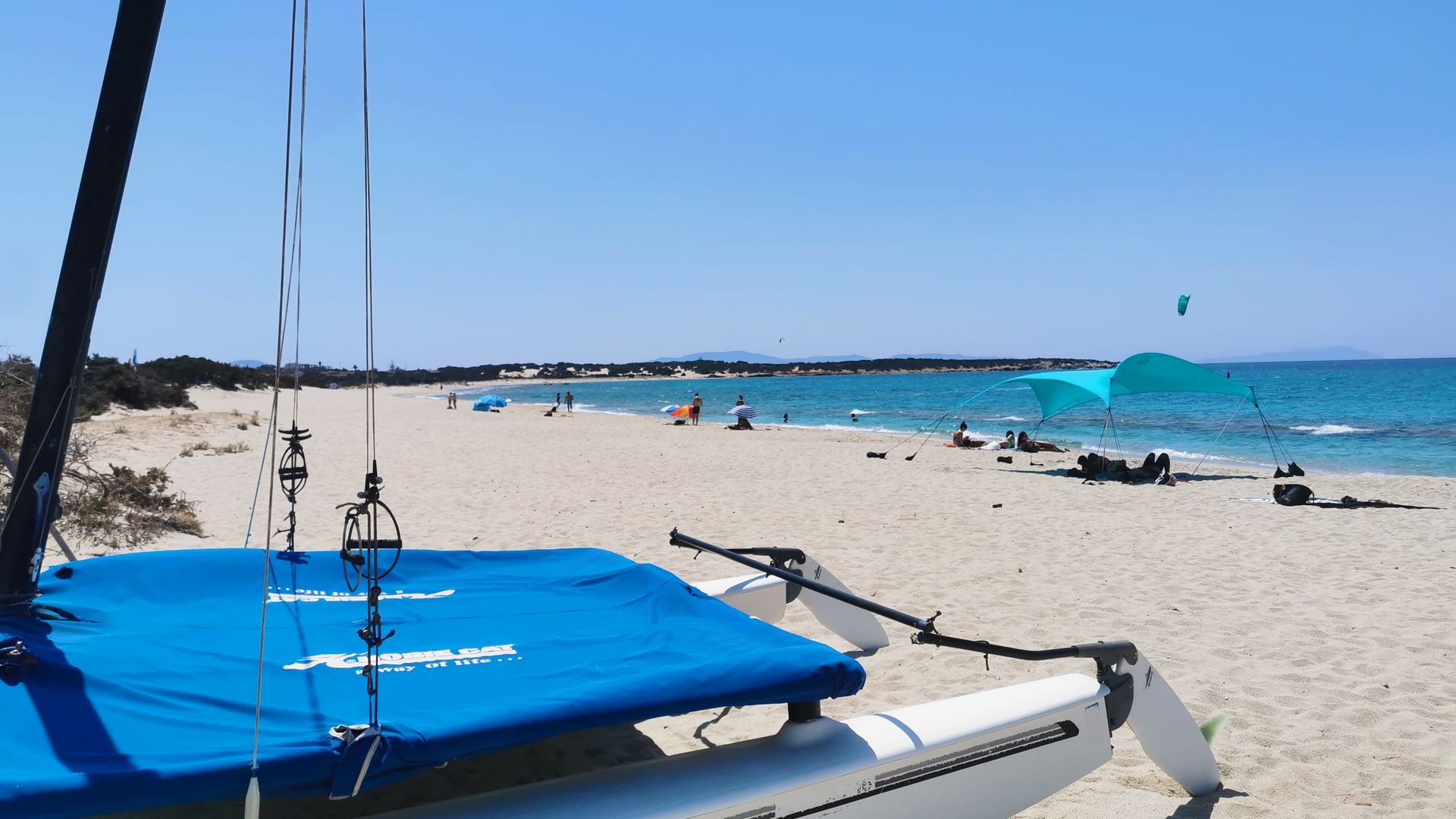 Paralia Glifada Kitesurfing in Naxos Greece with a catamaran boat in front on white sand, some people laying on the beach and a few kitesurfers surfing in the background