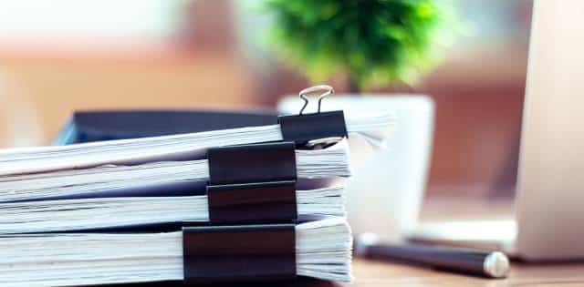 : A stack of business documents clipped together on a wooden desk in an office next to a plant, pen, and laptop.