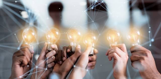 A group of hands holding up shining lightbulbs together in a row with digital overlays and a blurry background.