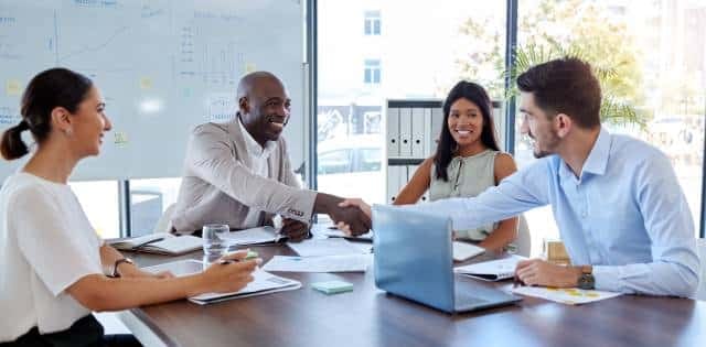 Four people sit at a table in a meeting room with documents in front of them. Two men shake hands while everyone smiles.