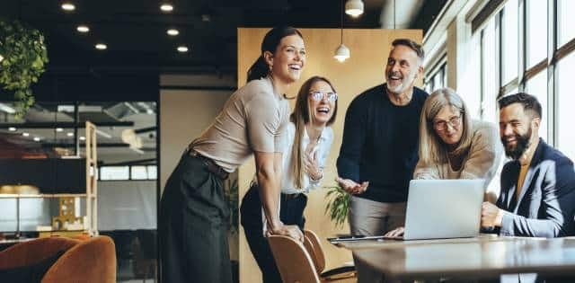 A small group of employees gathered around a table in the office. The employees are smiling and laughing with each other.