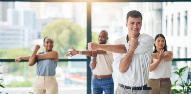 Four workers wearing business attire in an office exercise group stretching their arms. Behind them is a large window.