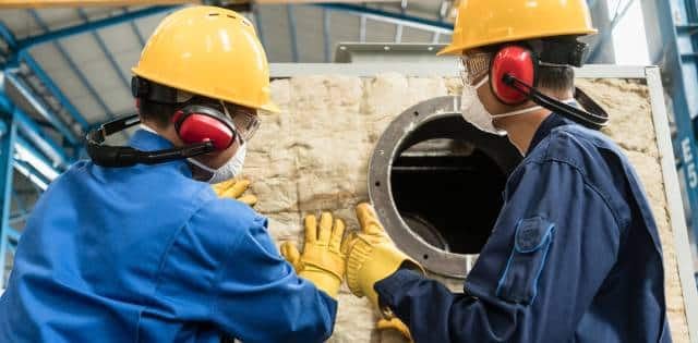 Two plant technicians applying industrial-grade insulation padding to boiler equipment, protecting it from heat loss.