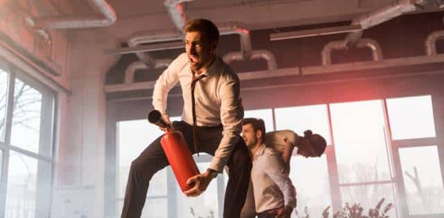 A scared worker standing on a desk, wielding a fire extinguisher in a smoke-filled office as two coworkers run by behind him.
