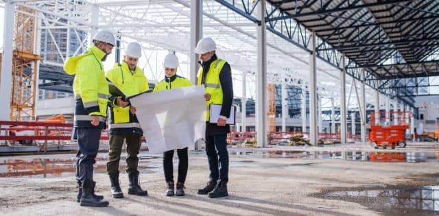 Four construction workers reading over their written construction plans while onsite for their building project.
