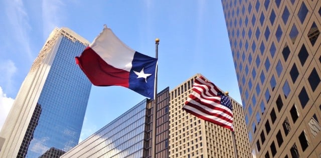 Flagpoles with the American and Texas flags flying in the wind in front of several skyscrapers in an urban city.
