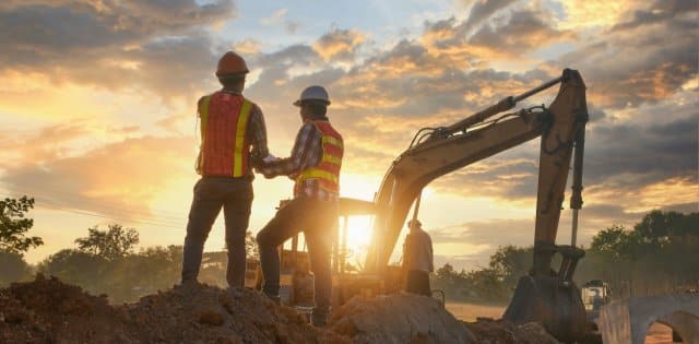 Two construction employees are working on the site and standing on a pile of dirt while the sun sets.
