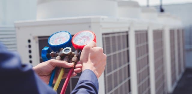 An HVAC technician is on a commercial roof holding a gauge with three cords next to large air conditioning units.