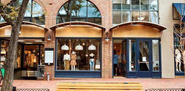 A row of warmly lit windows containing fashion mannequins with a doorway to the left, giving access to the clothing store.