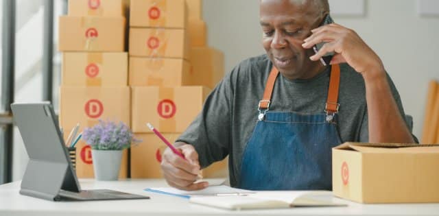 A man in a dark gray shirt and blue apron holds a phone to his ear while writing on a notepad. Boxes are behind him.