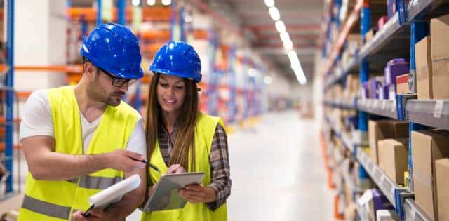 A man and a woman wearing yellow hi-vis vests and blue hard hats stand in a warehouse looking at clipboards.