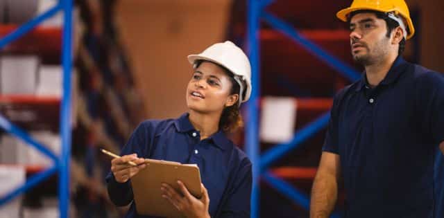 A man and a woman, both wearing navy polos and hard hats, glance at something beside them in a warehouse with racks in the background.