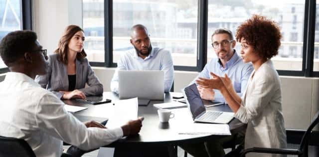 Five employees are talking around a conference table in an office. The table has laptops and papers.