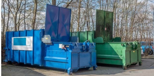 Two large trash compactors, one green and one blue, sit outside on the pavement in front of barren trees.