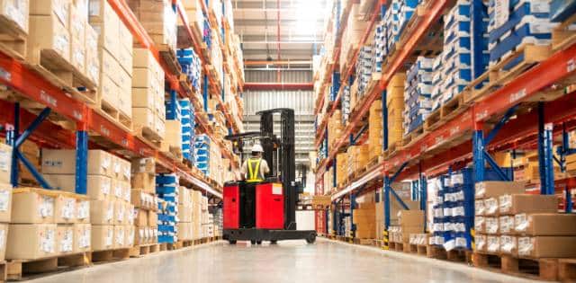 A warehouse worker driving a forklift between two industrial racks that are full of boxes stacked on pallets.