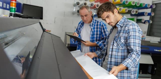 Two men look at a large-scale printer as one man guides the paper out of the machine while the other writes on a clipboard.