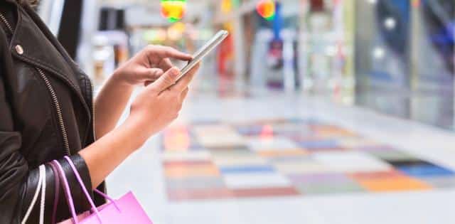 A side profile of a woman's torso as she stands in a shopping mall, carries three bags on her arm, and uses her phone.