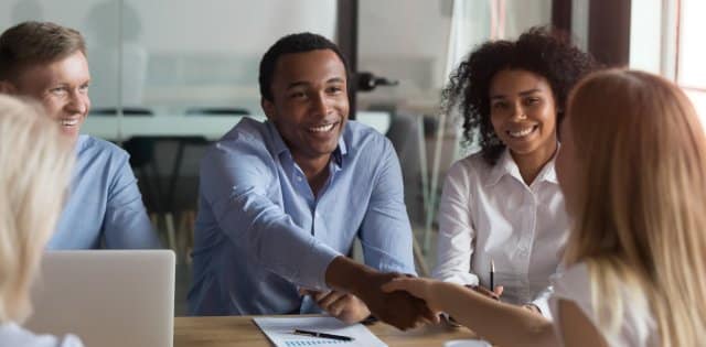 hree smiling people sit across a table from a young woman with long blonde hair as one man shakes her hand.