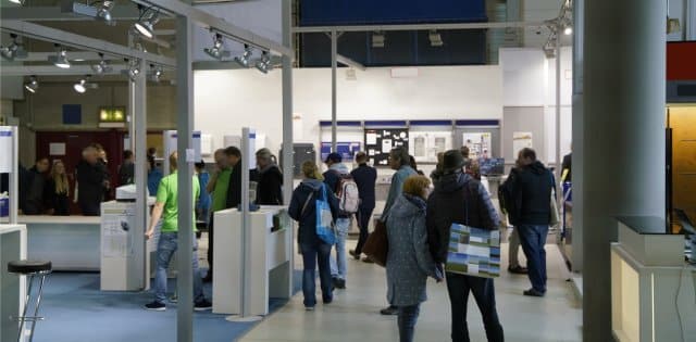 A crowded convention hall full of booths and people wandering around. A large projector screen hangs on the wall.