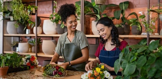 Two women smile as they wrap bouquets behind a flower shop counter with shelves of various pots behind them.
