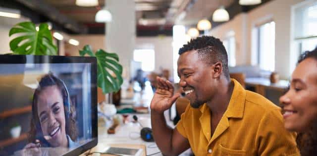 Employees in a hybrid office environment smile and wave at a woman on a video call wearing a headset.
