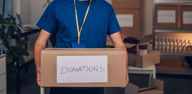 A smiling man with a beard and a laminate hanging from his neck carries a brown box labeled donations.