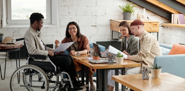 A young man in a wheelchair holds a large piece of paper while talking to three other people sitting around a wooden desk.