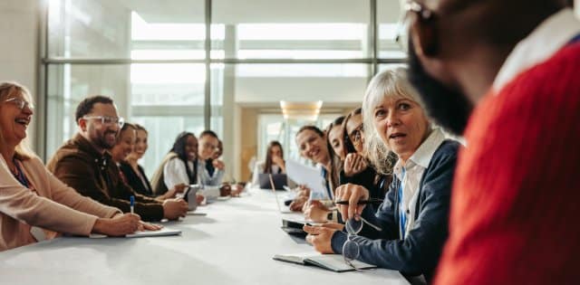 A large group of professionals sit at a conference table and collaborate in a workspace with glass partition walls.