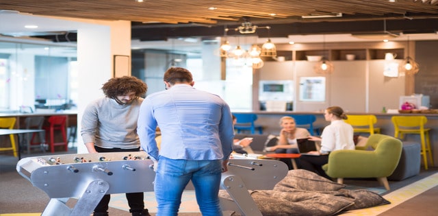 A modern office space. Two employees play foosball in the foreground while two employees behind them sit on a couch and chat.