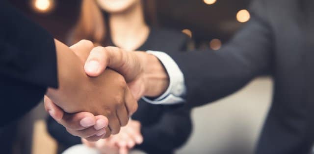 A close-up of two people in business attire shaking hands while sitting down. A woman smiles in the background.