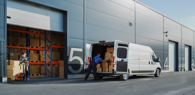 A white delivery truck parked outside an open garage to a warehouse. Two workers load the truck with packages.
