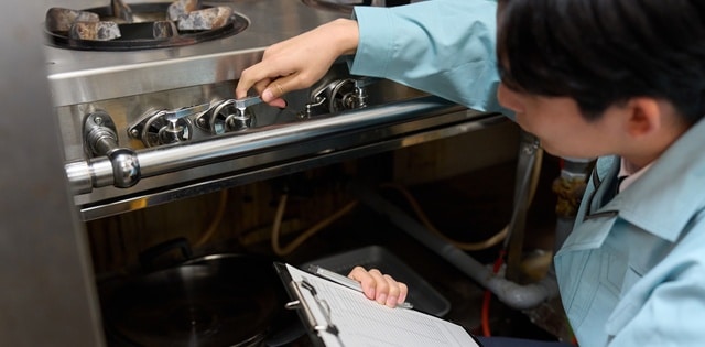 A person in a blue jacket holding a clipboard and pen kneels in front of a commercial stovetop to inspect it.