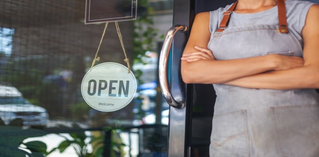 A woman wearing an apron stands with her arms crossed leaning on a restaurant door with an open sign hanging in the window.