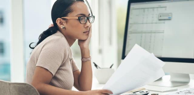 A person sitting at a desk looks at sheets of paper while a nearby computer monitor displays a spreadsheet.
