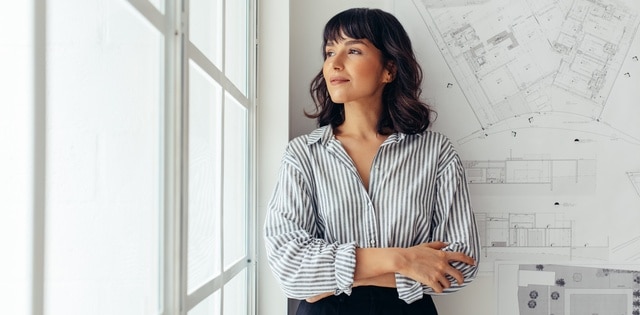 A woman in a striped shirt stands proudly with her arms crossed in front of a blueprint and next to a window in an office.