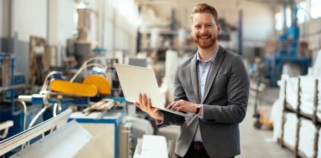 A man wearing a gray blazer and black slacks holding a laptop on a manufacturing floor checking machines.