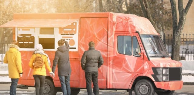 Four customers in black and yellow coats stand outside a red food truck with a geometric design on a city street with snow.