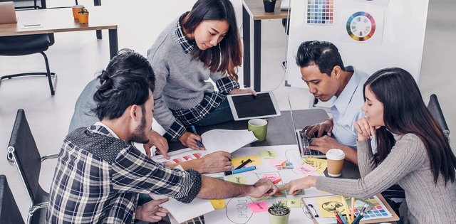 A group of five teammates collaborating at a work table with a tablet, laptop, and color palette to review creative branding.