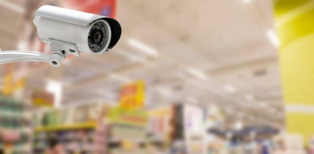 A large, well-lit retail store with fully stocked shelves. In the foreground is a white security camera pointed at an angle.