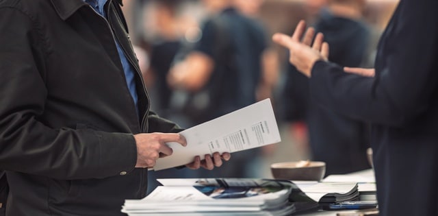 Two people stand across from one another at a tradeshow table. One person holds a piece of paper in their hands.