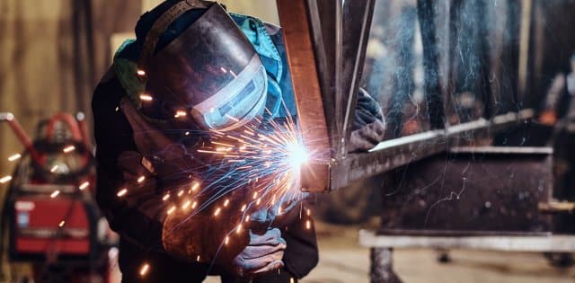 A man wearing a protective face mask and coveralls using a welder to fuse two pieces of metal together.
