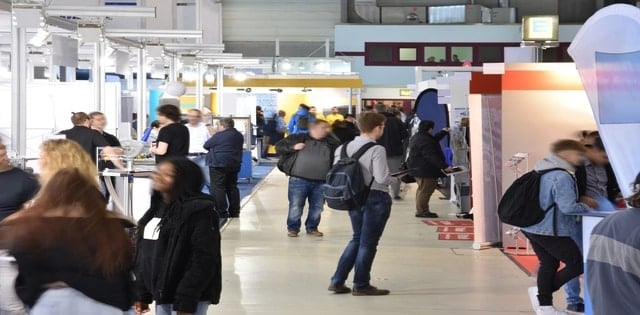 A crowd of event attendees explores several booths at a convention center, mingling with businesses and guests.