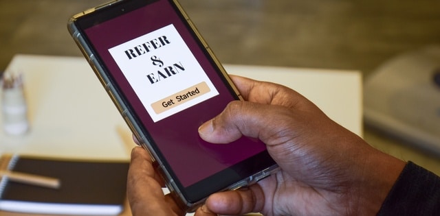 A man holding a smartphone with a maroon screen and a text display reading "refer & earn" above a button reading "get started."