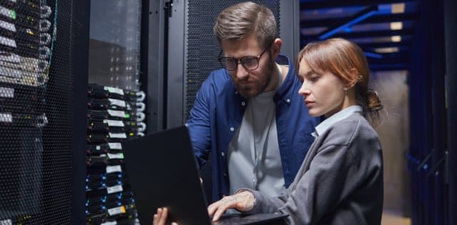 Two IT professionals reviewing data on a laptop between rows of black server towers in a data center.