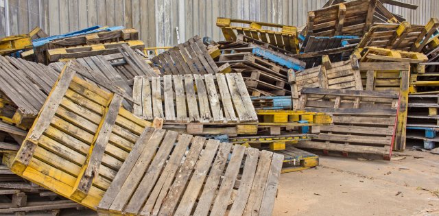 Dozens of dirty and broken wooden pallets are piled haphazardly on the ground next to a large building.