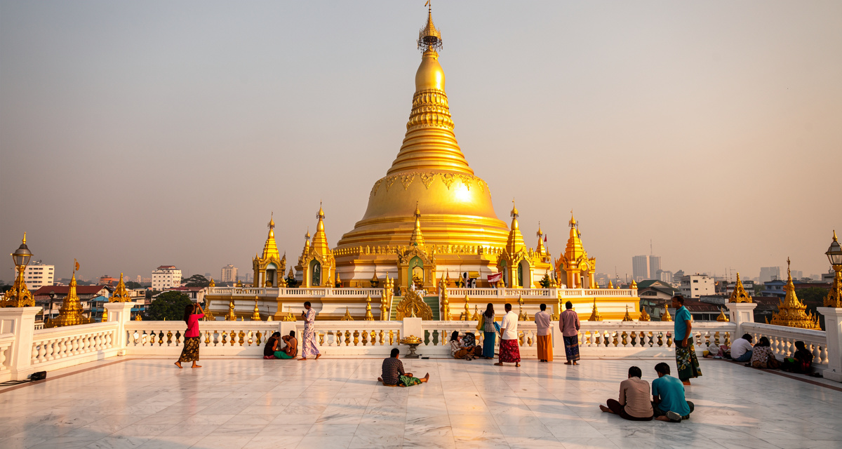 Shwedagon Pagoda golden stupa towering over Yangon at golden hour with its massive gilded dome catching warm sunlight, devotees walking around the white marble platform, representing Myanmar and the +95 international dialing code