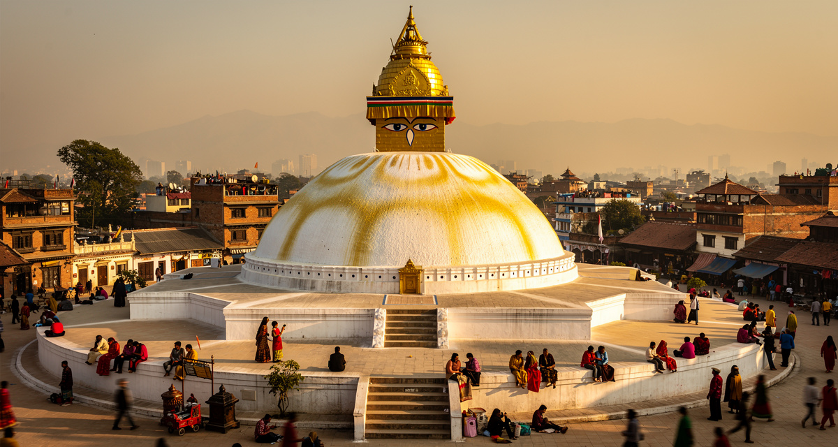 Boudhanath Stupa in Kathmandu at golden hour with its massive white dome and golden spire draped in colorful prayer flags, representing Nepal and the +977 international dialing code