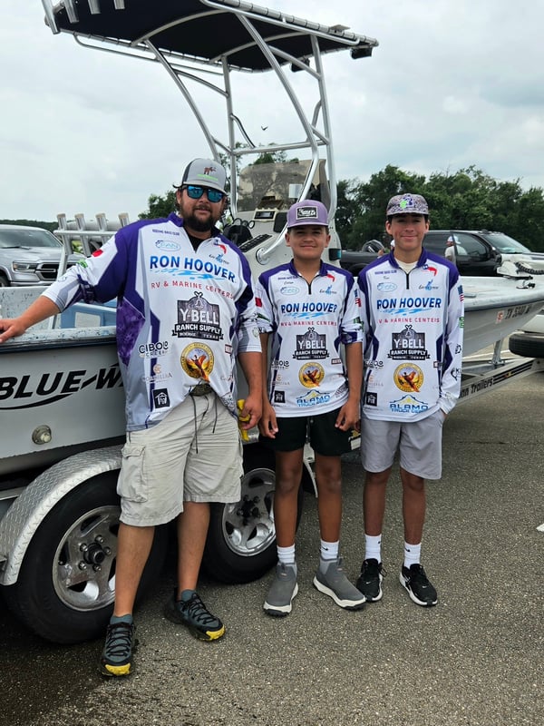 Three youth anglers in fishing jerseys pose by their boat.