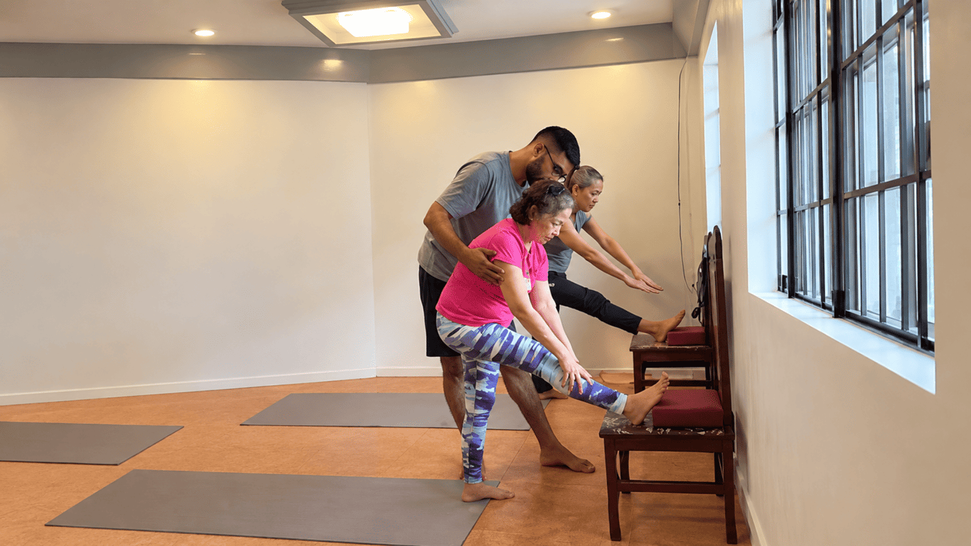Teacher Kirti assists a student with a stretching pose emphasizing proper alignment and individualized guidance during practice.
