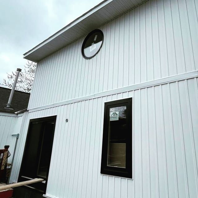 Black round Panes window in a white-sided gable above a rectangular window and patio opening, shared by Renaud Construction